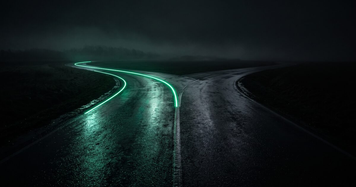 Estrada molhada à noite a bifurcar em duas vias, uma delas traçada por uma linha de luz verde: a escolha entre Meta Ads e Google Ads