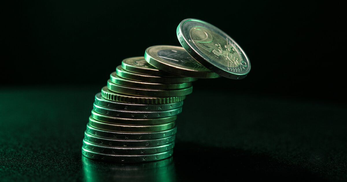 Pilha de moedas de euro com uma moeda a tombar no topo, iluminada a verde sobre fundo escuro: o custo de anunciar no Facebook em Portugal